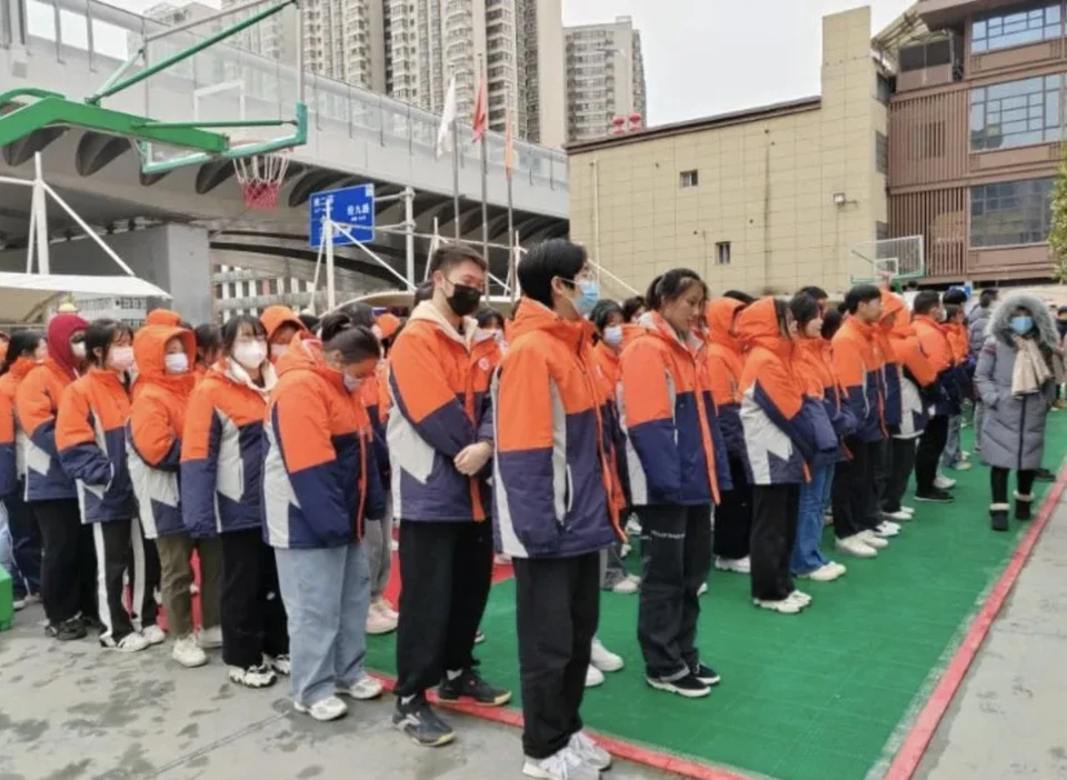 补习学校学生在操场列队集合，身穿橙色与深蓝拼色校服，背景为教学楼、篮球架与高层住宅