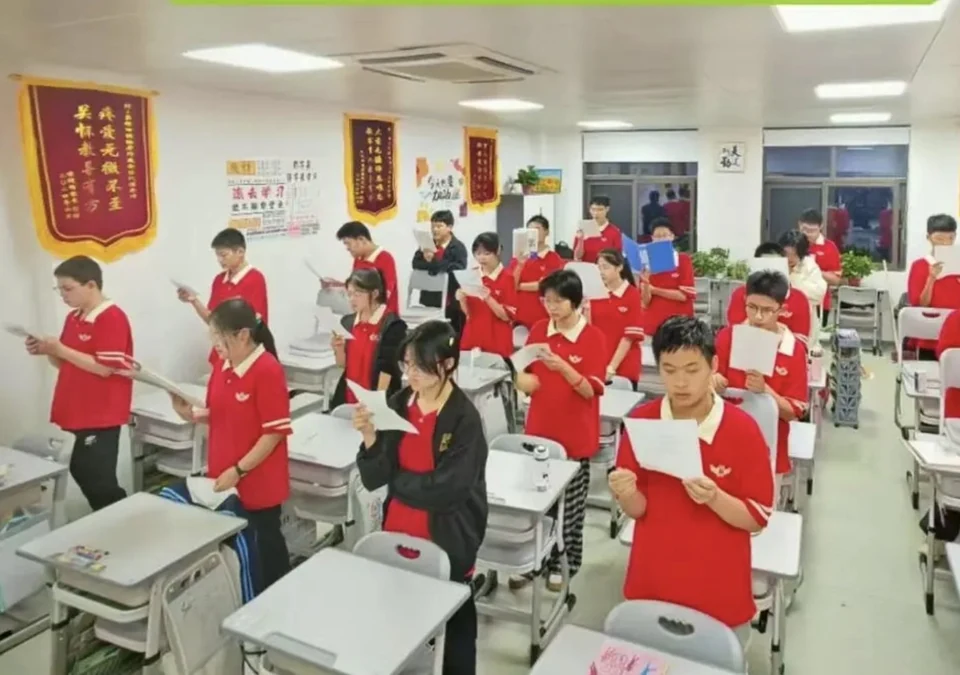 教室内穿红色校服的学生集体站立朗读背诵，教室墙面挂有多面荣誉锦旗与学习标语
