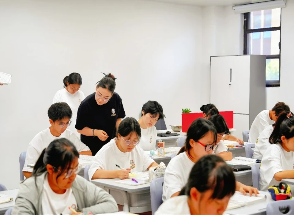 小班教室内学生穿白色校服做题，一位老师站在学生座位旁俯身进行个别指导