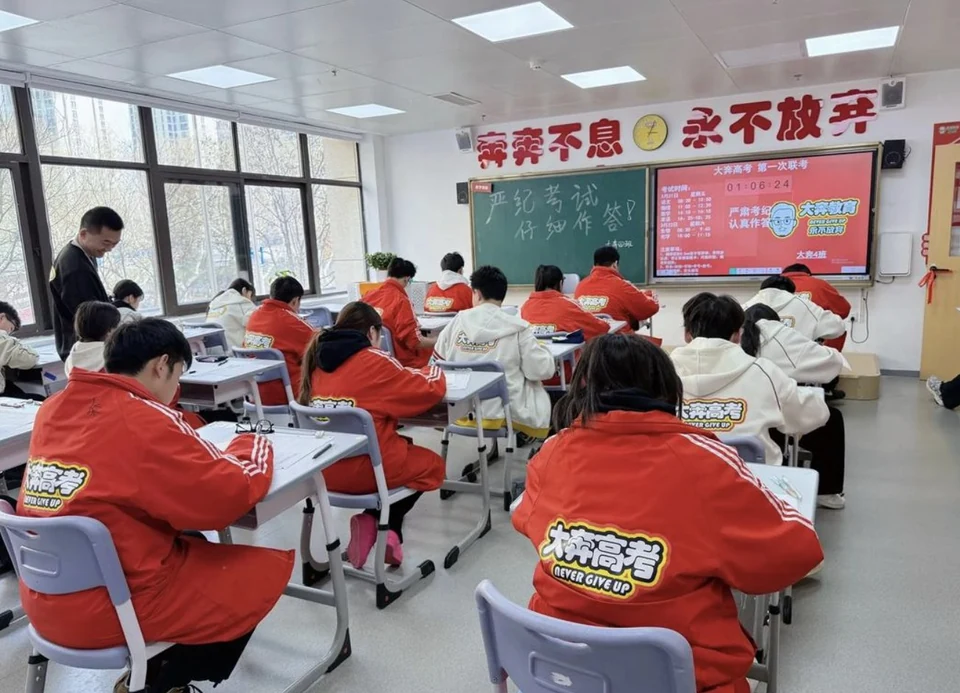 教室内学生穿红色校服参加联考，后墙屏幕显示「大奔高考 第一次联考」的场次与班级信息，黑板上写着严纪考试仔细作答