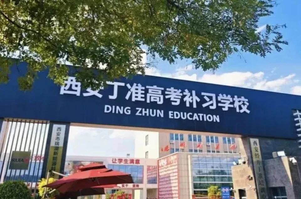 西安丁准高考补习学校校门实拍，深蓝色门头招牌上写有校名与英文名，门前有绿树与红色遮阳伞