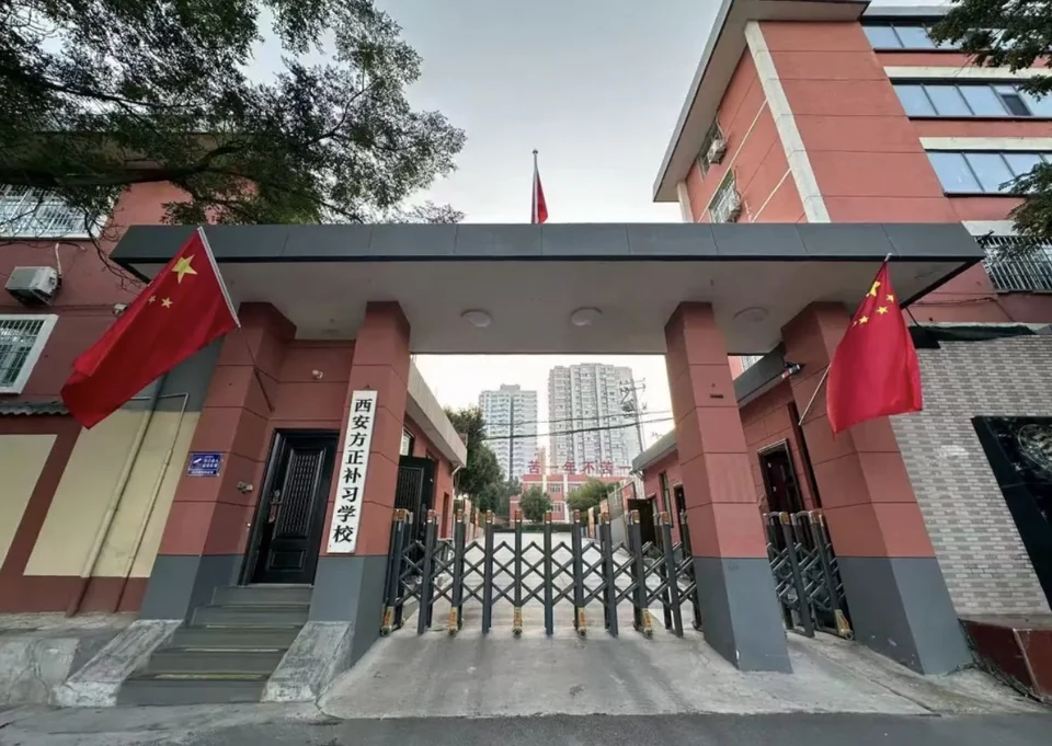 西安方正补习学校校门实拍，红棕色门柱上挂有写着校名的白色立牌，两侧悬挂国旗，门前是灰白台阶与黑色伸缩门