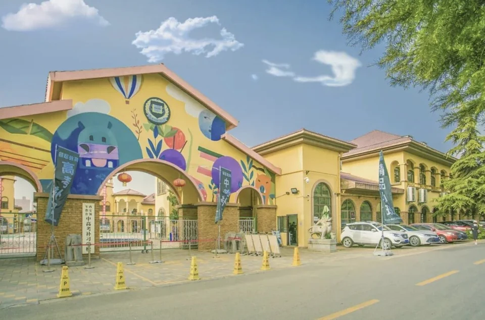 西安领军教育全日制封闭校区校门实景，黄色主楼带彩绘墙面，门口立有中高考补习标牌与高考冲刺、中考补习横幅