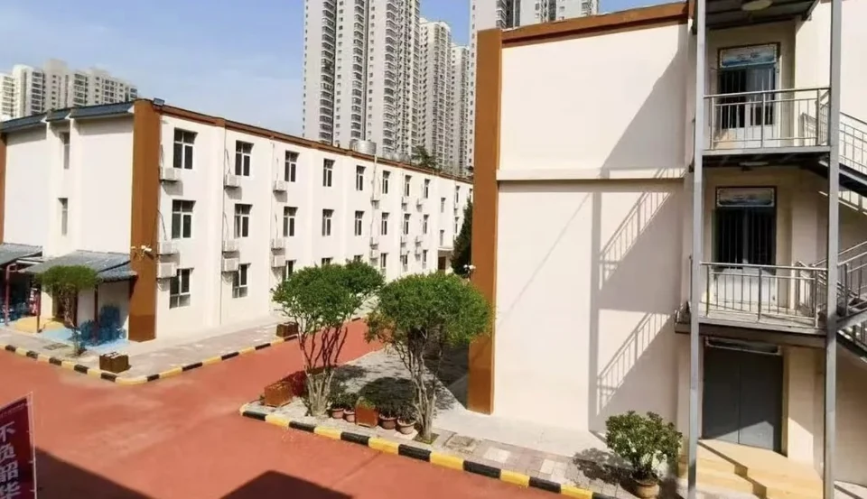 补习学校校园实景，白色多层教学楼配橙色装饰线条，楼前为红色地面与绿化，右侧有室外钢梯，远处可见高层住宅楼